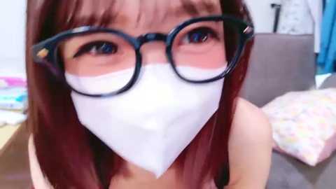 Media: A video of an Asian woman with straight brown hair and glasses, wearing a white surgical mask, sitting on a grey couch with colorful pillows in a living room.