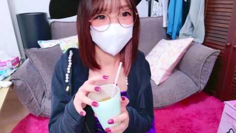 Media: A video of an East Asian woman with straight brown hair and round glasses, wearing a white face mask, black cardigan, and pink nail polish, holding a drink in a room with a pink rug, grey couch, and clothes hanging.