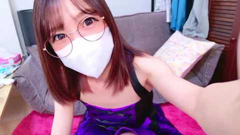 Media: Video of an East Asian woman with straight brown hair, wearing round glasses and a white surgical mask, seated on a pink blanket in a cozy living room.