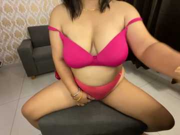 Media: Video of a plus-size woman with medium skin tone, wearing a bright pink bra and panties, seated on a gray chair, in a room with patterned wallpaper and a dark curtain.