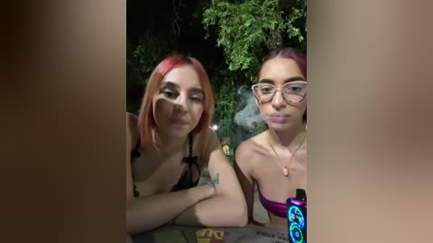 Media: Video of two young women with light skin, one with red hair and black top, the other with brown hair and glasses, smoking weed, outdoors at night.