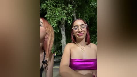 Media: Video of two young women outdoors, one with red hair and glasses, wearing a metallic pink bandeau top, the other with a black top, in a lush green forest background.