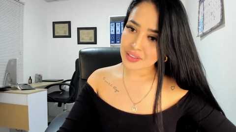 Media: Video of a Latina woman with long black hair, light skin, wearing a black off-shoulder top, sitting in an office chair, tattoos on her shoulders, framed certificates on the wall, desk with a computer monitor.