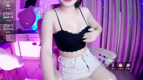 Media: Video of a young Asian woman in a black lace crop top and high-waisted white shorts, sitting on a white chair. Background features a pink-lit room with a vanity table and curtains.