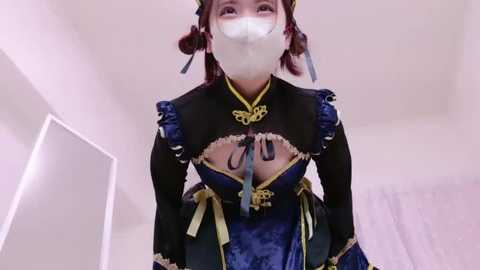 Media: Video of an East Asian woman in a blue and gold Chinese dress with ruffled sleeves, face mask, and pigtails, indoors with white walls and a window.