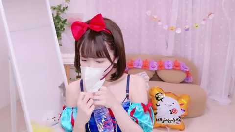 Media: A video of a young girl with straight black hair, a red bow, and a white face mask, holding a phone, in a pink room with a bed, a stuffed toy, and a curtain.