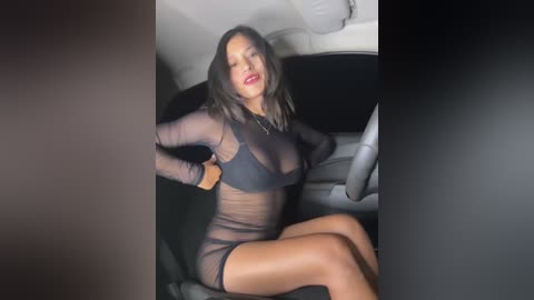 Media: Video of a Latina woman with medium brown skin and long dark hair, wearing a sheer black mesh top and matching shorts, sitting in a car, her legs crossed, lips slightly parted.