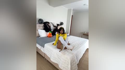 Media: Video of a woman with medium skin tone and long dark hair, dressed in a yellow jumpsuit and white knee-high socks, sitting on a white bed with a grey headboard, in a minimalist, bright bedroom.