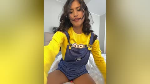 Media: Video of a woman in a Minnie Mouse costume with a yellow shirt, blue overalls, and oversized eyes, sitting on a bed in a bright, modern room.