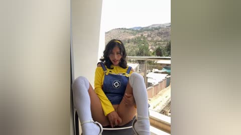 Media: A video of a young woman with medium skin tone and dark hair in a yellow and blue superhero costume, sitting on a balcony with a scenic mountainous background.