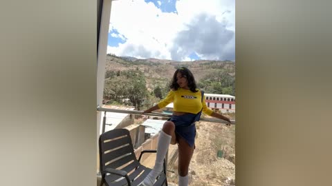 Media: Video of a curvy woman with long black hair, wearing a yellow crop top, blue denim shorts, and white knee-high socks, posing on a balcony overlooking a mountainous landscape with a partly cloudy sky.