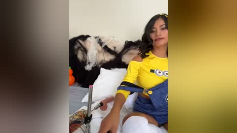 Media: A video features a young woman with dark hair, wearing a yellow and blue soccer jersey, white thigh-high socks, and denim overalls, sitting on a bed with a black and white dog.