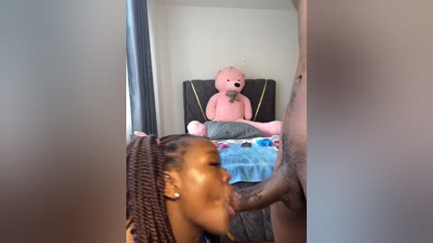 Media: A video shows a young Black woman with braided hair performing oral sex on a naked Black man, while a pink teddy bear sits on a bed in the background.