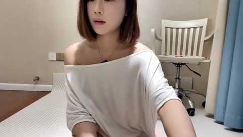 Media: Video of a young Asian woman with shoulder-length brown hair, wearing a white off-shoulder top, sitting on a mat in a sparsely furnished room with a white chair and blue curtain.