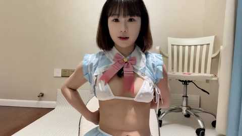 Media: A video of an Asian woman in a revealing maid costume with a pink bow, sitting on a white mat in a beige room with a white chair and a computer desk.