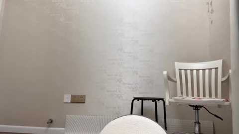 Media: Video of a small, sparsely furnished room with a white-painted brick wall, a white chair, a small stool, and a bed with white bedding in the foreground. The room is minimally decorated with a single electrical outlet on the wall.