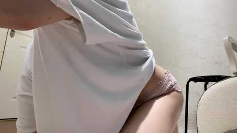 Media: Video of a woman lifting her white shirt to reveal pink lace panties, in a minimalist room with a beige wall and a black chair.