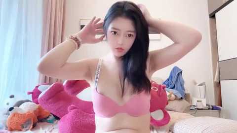 Media: Video of a young East Asian woman with long black hair, wearing a pink bra, standing in a messy bedroom with plush toys, a bed, and a dresser in the background.