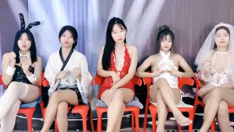 Media: Video of five Asian women in provocative bunny costumes, sitting on red plastic chairs, wearing lingerie, makeup, and wigs, against a backdrop with white and grey drapes.