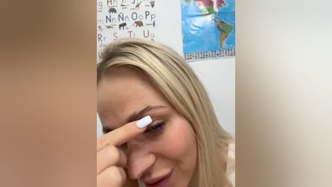 Media: Video of a blonde woman with light skin, applying makeup to her left eye with a white eyeliner pencil. Background features a colorful world map and a decorative wall hanging.