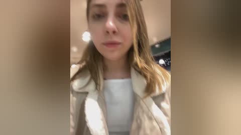 Media: A video of a young woman with light skin and shoulder-length brown hair, wearing a white shirt and beige coat, standing indoors with blurred background.