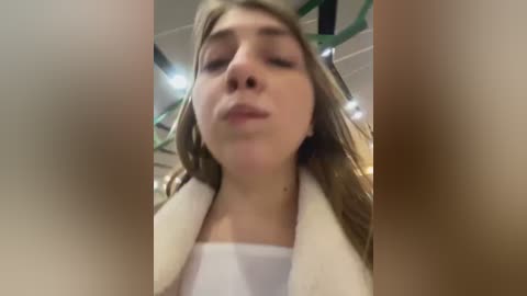 Media: Video of a young Caucasian woman with fair skin, long brown hair, wearing a white top and beige jacket, captured from a low angle, looking upward with a neutral expression.