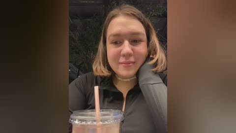 Media: A candid video of a young woman with shoulder-length blonde hair, wearing a black jacket, holding a colorful plastic cup with a pink straw, sitting outdoors against a blurred green background.