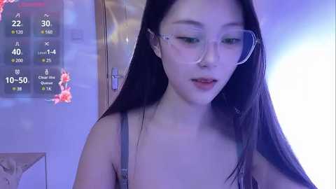 Media: A video of an Asian woman with long black hair, wearing glasses and a strapless top, indoors with a digital clock and a colorful background.