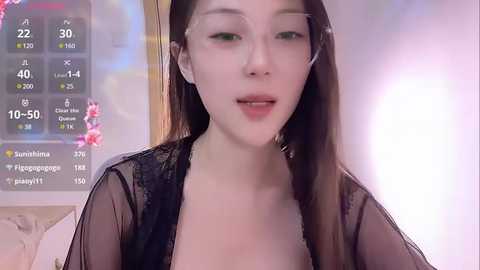 Media: A video of an East Asian woman with fair skin, long straight black hair, and green eyes, wearing a sheer black lace robe, standing in front of a digital weather display showing 10\u00b0C.