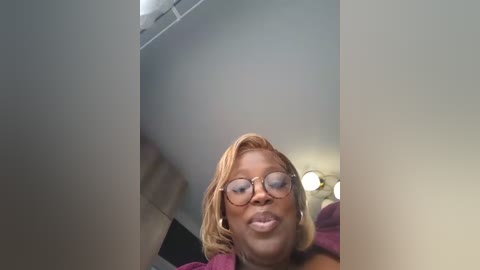 Media: Video of a Black woman with short, straightened hair, wearing glasses and a maroon top, looking up in a dimly lit room with white walls and a ceiling fan.