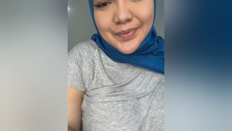 Media: Video of a smiling woman with light skin and dark hair, wearing a blue hijab and a grey short-sleeved t-shirt. The background is a soft gradient of blue to white.