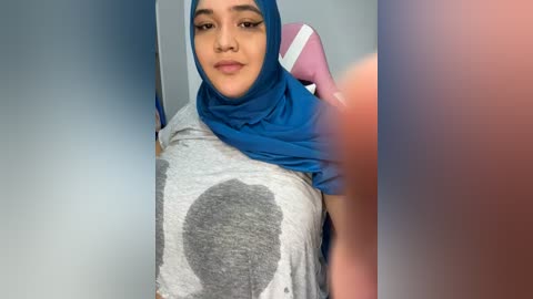 Media: Video of a young woman with medium skin tone and dark hair, wearing a blue hijab and gray shirt with a large, abstract gray design, standing indoors with blurred background.