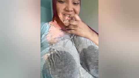 Media: Video of a smiling woman with medium skin tone, wearing a grey sweatshirt, her face and hands covered in pink Holi powder. She is indoors with a blurred background.