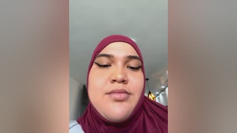 Media: Video of a young woman with light brown skin, wearing a maroon hijab and light makeup. She has a neutral expression, with her eyes closed and lips slightly parted. Background shows a modern, well-lit room with a white ceiling and visible furniture.