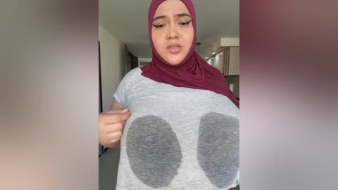 Media: Video of a young woman with medium brown skin and a maroon hijab, wearing a grey t-shirt with large grey circles over her breasts, taken indoors.