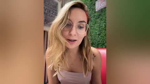 Media: Video of a blonde woman with fair skin, wearing glasses and a white strapless top, looking surprised while seated. Background features green wall and red furniture.