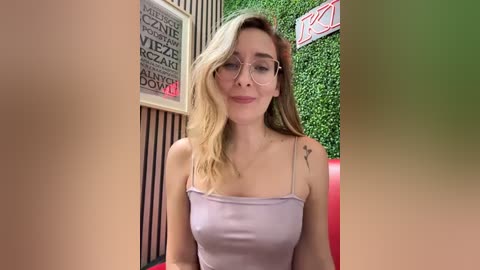 Media: A video of a young woman with fair skin, shoulder-length wavy blonde hair, and glasses, wearing a light purple spaghetti-strap top, sitting in a red booth with a green wall background.