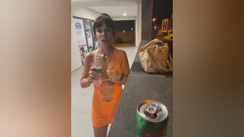 Media: Video of a woman with dark hair and a slender physique, wearing a tight, orange dress, holding a green drink can, standing in a dimly lit parking lot with a large brown bag and a green and red can in the background.