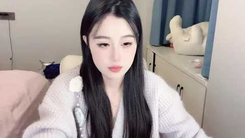 Media: A video of a young Asian woman with long, straight black hair and fair skin, wearing a light grey robe, sitting on a bed with a pink blanket. The room has a white dresser and a blue curtain in the background.