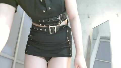 Media: Video of a fair-skinned woman in a black, button-up crop top and a short black leather skirt with silver studs and belt, standing in a brightly lit room with white walls and a mirror.