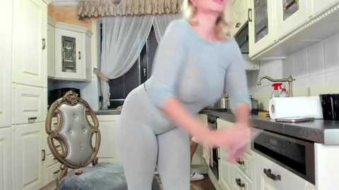 Media: A blurred video of a woman in a light blue, long-sleeved jumpsuit bending over in a kitchen with white cabinets, wooden floors, and a tufted chair.