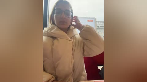 Media: Video of a blonde woman with glasses, wearing a beige hoodie, adjusting her hair near a large window, with blurred background of an office building.