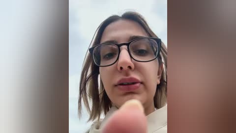 Media: A video of a young woman with straight brown hair, wearing black-framed glasses, and a beige jacket, captured from a low angle, emphasizing her direct gaze.