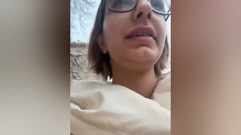 Media: Video of a light-skinned woman with short brown hair, wearing glasses, and a beige top, partially obscured by a blurred vertical object. Background shows a stone wall.