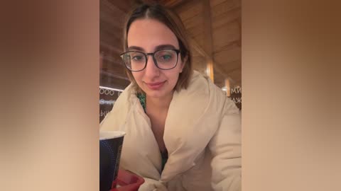 Media: A video of a young woman with fair skin, glasses, and shoulder-length brown hair, wearing a beige robe, seated in a wooden interior with a dark blue cup in front of her.