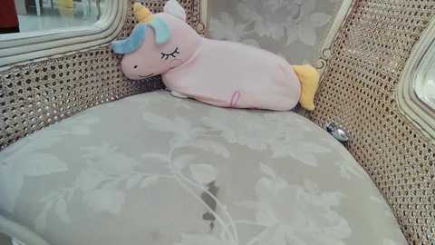 Media: Video of a vintage wicker chair with a pastel pink unicorn pillow and floral-patterned cushion. The chair has intricate detailing and a soft, light beige fabric.