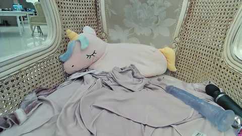 Media: Video of a plush unicorn toy with closed eyes, lying on a light grey bedsheet, surrounded by a beige wicker chair and a blue vibrator, in a plush, floral-patterned room.