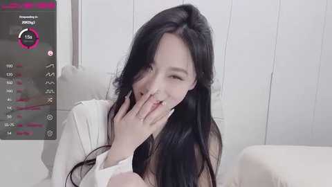 Media: Video of a young East Asian woman with long black hair, wearing a white shirt, covering her mouth with her hand, and looking embarrassed. Background shows a white wall and a smartphone screen displaying \"Xiaoyu\" and temperature data.