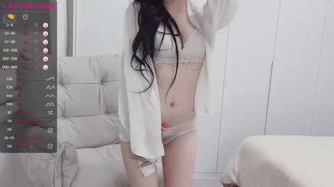 Media: Video of a slender, fair-skinned woman with long black hair, wearing light pink lingerie, standing in a minimalist white bedroom.