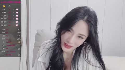 Media: A video of an East Asian woman with long black hair, fair skin, and a slender build, wearing a white shirt, smiling in an indoor setting with a digital clock displaying time.